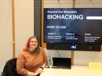 Abi Farnsworth sits beside a large monitor displaying the project, "Beyond the Wearable: Biohacking."